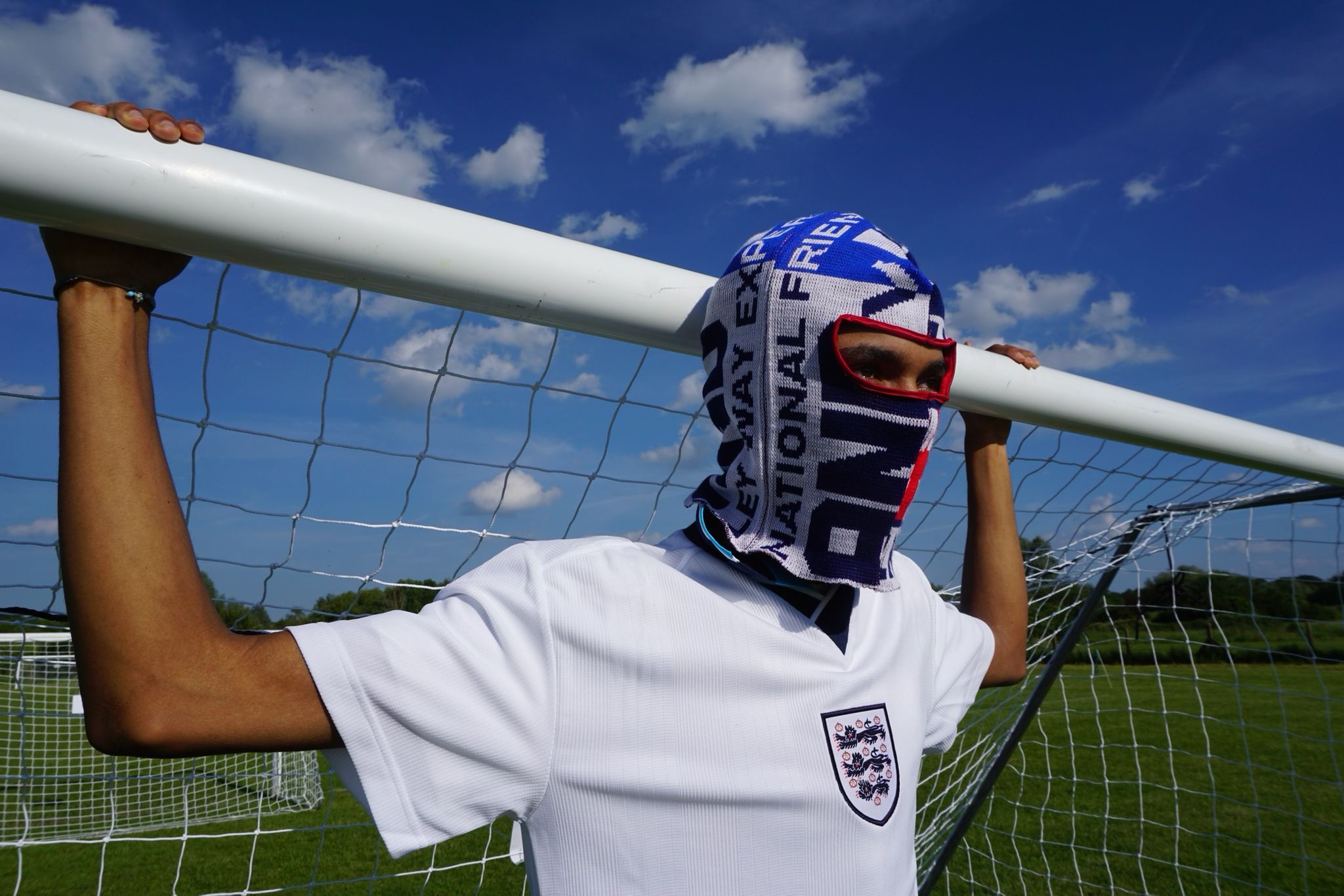 Our model wearing our England football scarf balaclava with a classic white England shirt leaning against a football goal